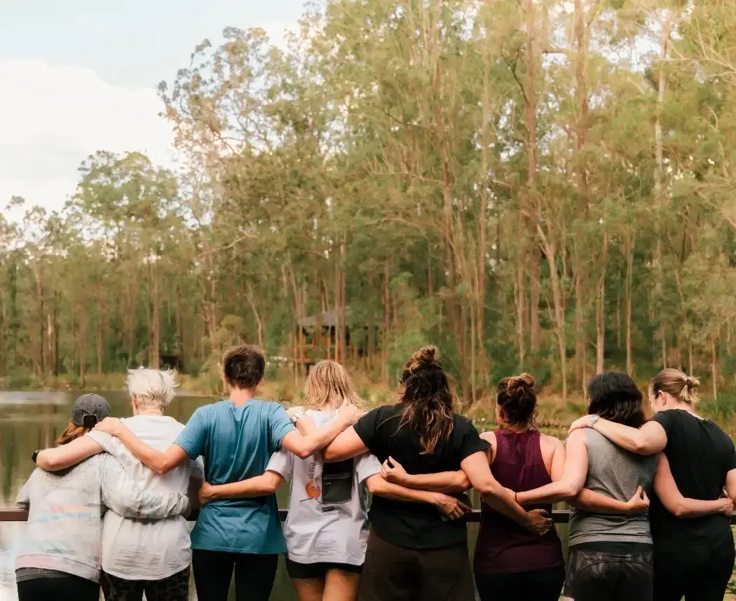 Group of friends with arms around each other, facing a lake in a forested setting, conveying companionship and unity.