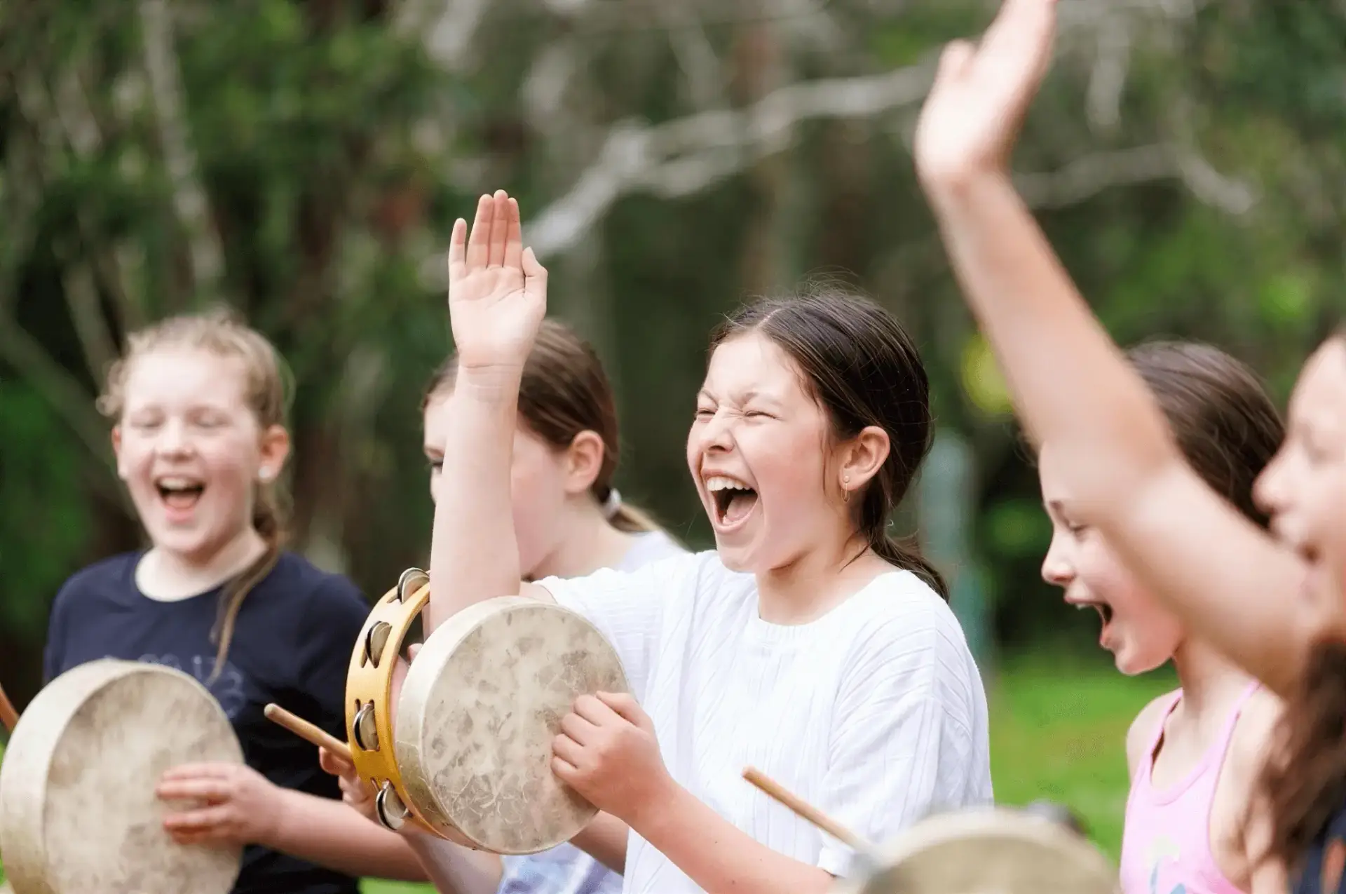 Group of young people playing handheld drums outdoors, raising their hands in a lively beat.